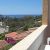 Une vue calme depuis un balcon, avec des maisons, des montagnes et la mer au loin sous un ciel bleu.